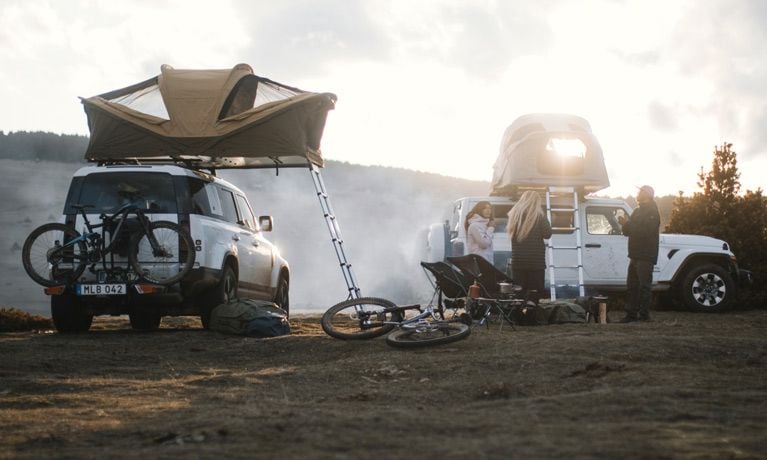 Two vehicles with Thule rooftop tents are parked in a field where people have their camp set up.