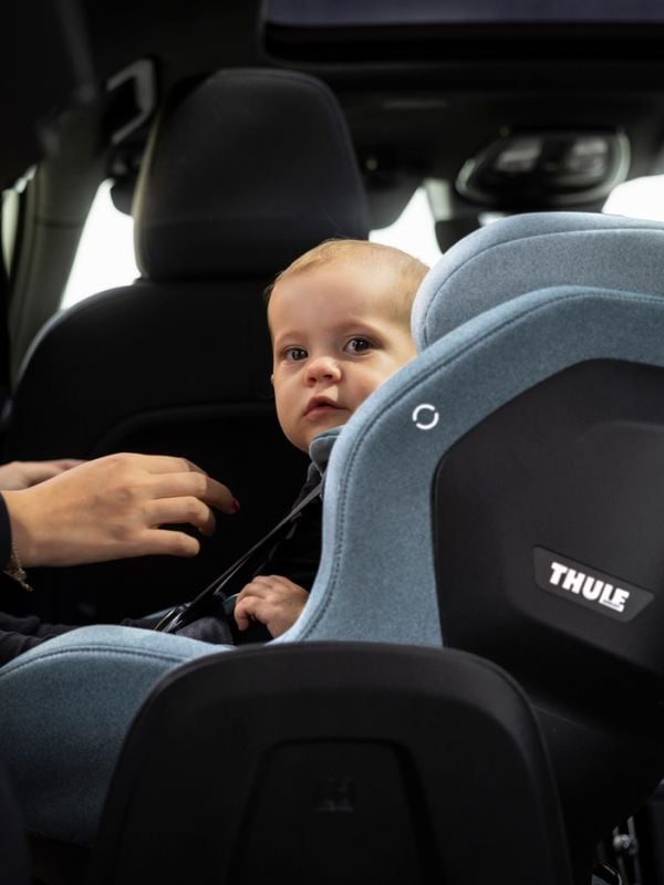 A young toddler sitting in a Thule Elm toddler car seat in light grey. A person's hands are near the toddler's straps.